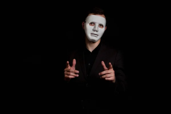 Siyah bir arka plan beyaz maskeli bir adam duruyor. Bir gest yapar