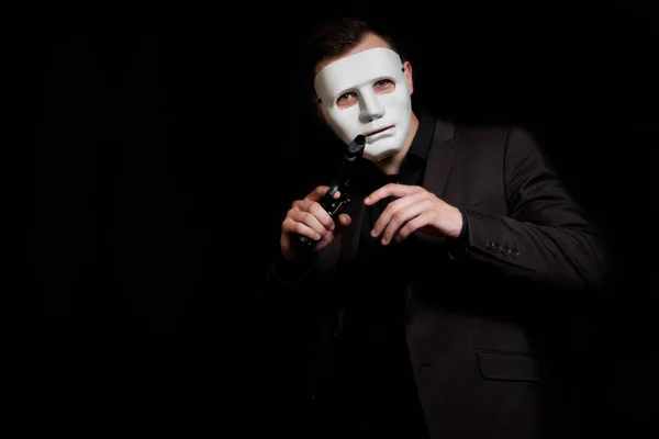 A man in a white mask on a black background. He holds the gun in