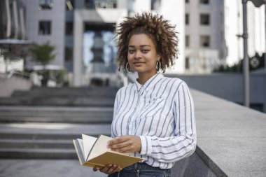 Koyu tenli kız bir kitapla sokakta duruyor. O afro Amerikan ofis binası yakınında ve düz görünüyor.