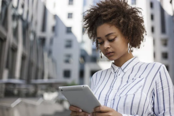 Afro american girl is standing on the street near the office building and holds a laptop in her hands. She works on a tablet.