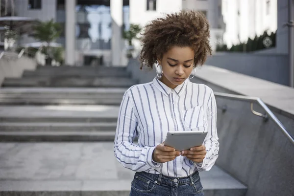 A dark-skinned girl stands on the street near an office building. It works on a tablet.