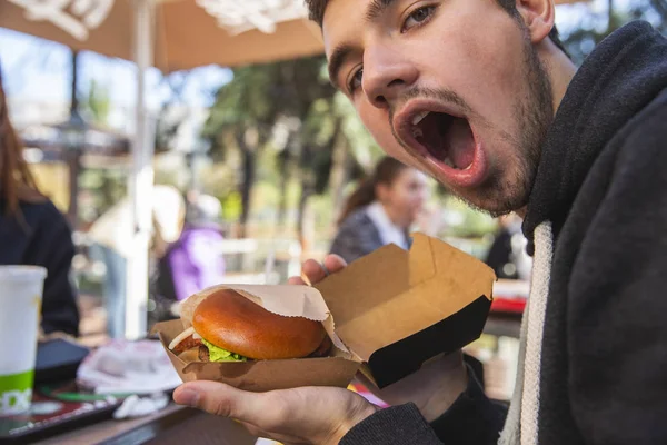 Satisfied young man looking at the camera, his mouth is wide open. He is holding burger in his hands. — Stock Photo, Image