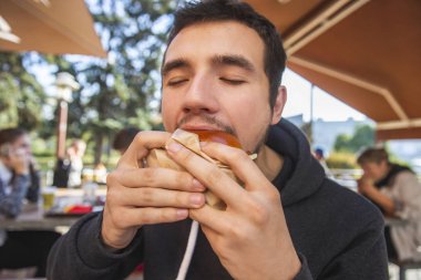 Genç adam, restoranda terasta otururken onun burger iştahla yiyor. Gözleri kapalı.