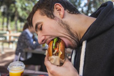 Young man is greedily eating his burger while sitting at the restauraunt terrace. His mouth is wide open. clipart