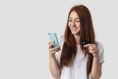Gri arka planda beyaz t-shirt üzerinde izole kırmızı saçlı kız. O telefon ve kredi kartı elinde tutar. Mağaza, Sigara nakit ödeme ve online alışveriş alışveriş kavramı