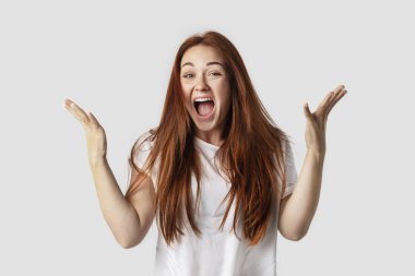 Gri arka planda beyaz t-shirt üzerinde izole kırmızı saçlı kız. O ellerini kaldırdı ve sevinir. Duygusal bir dalgalanma sevinç kavramı