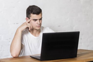 Genç adam, düşünceli dizüstü bilgisayar ekranında benziyor, dizüstü bilgisayarda çalışır ve masada oturur.