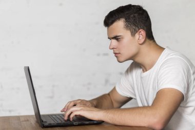 Masada oturan ve bir dizüstü bilgisayar üzerinde çalışan ciddi genç adam.