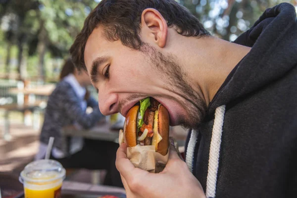 El joven está comiendo codiciosamente su hamburguesa mientras está sentado en la terraza del restaurante. Su boca está abierta. . — Foto de Stock