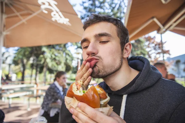 Young man wipes his mouth with his right hand at satisfaction, his eyes are closed. He is holding bitten hamburger at his left hand. Tasty food concept. — Stock Photo, Image