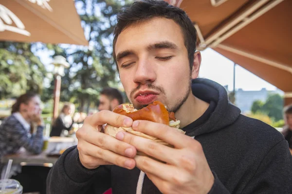 Genç adam onun ısırıldı burger tutuyor memnun, gözleri kapalı. O fastfood Terrace'da oturuyor.