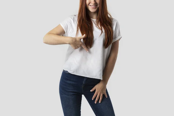 Giovane ragazza dai capelli rossi isolata su sfondo grigio in t-shirt bianca. Lei punta il dito verso il basso — Foto Stock