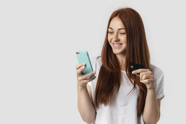 Joven chica pelirroja aislada sobre fondo gris en camiseta blanca. Ella sostiene el teléfono y la tarjeta de crédito en su mano. El concepto de compras en la tienda, pagos sin efectivo y compras en línea — Foto de Stock