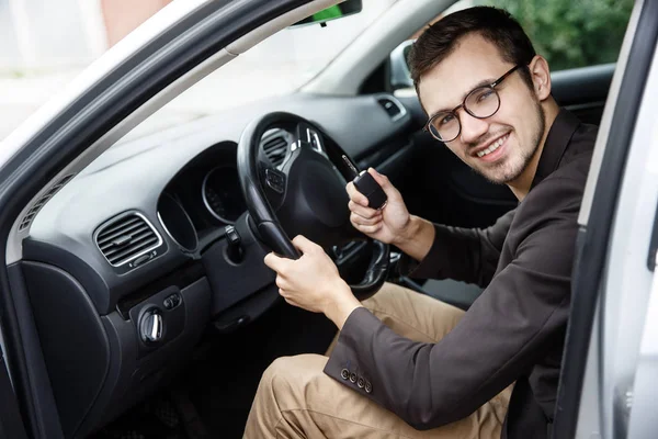 Tevreden jonge bestuurder zit aan zijn auto tijdens het kijken naar de camera. Hij houdt de sleutels aan zijn rechterhand. Zijn linkerhand is op het stuurwiel. — Stockfoto