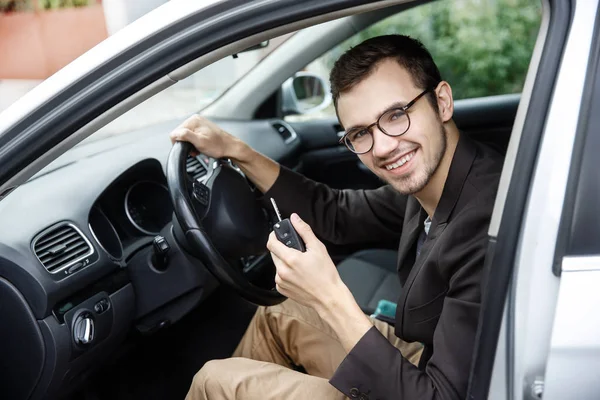 Contented young driver is sitting at his car while looking at the camera. He is holding the keys at his left hand. His right hand is on the steering wheel.