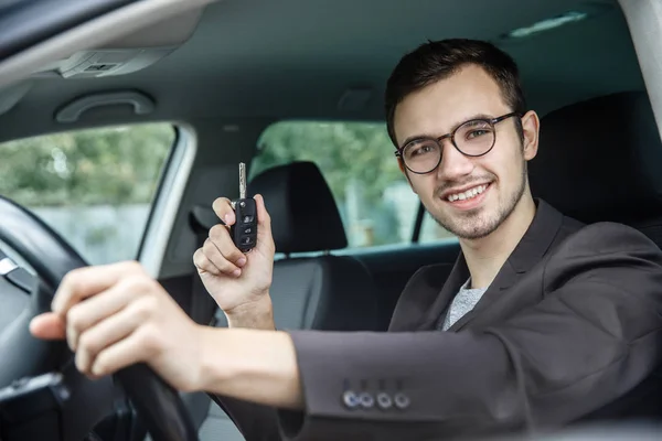Szczęśliwy młody facet siedzi w jego samochód patrząc na kamery. W prawej ręce trzyma klucze. Jego lewa ręka jest na kole kierownicy. — Zdjęcie stockowe