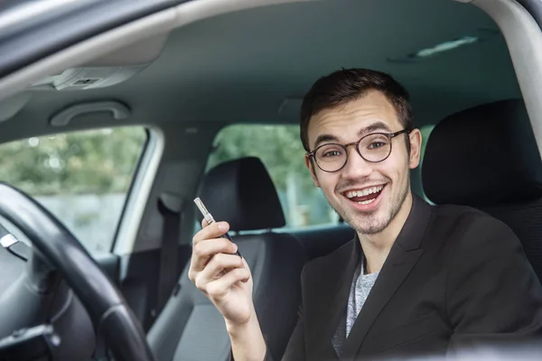 Tevreden jonge man houdt de sleutels aan zijn rechterhand tijdens het kijken naar de camera. Hij zit bij zijn auto. — Stockfoto