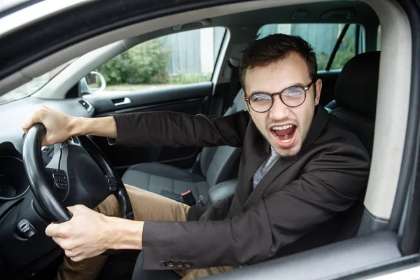 Woedend jonge man is kijken naar de camera tijdens de vergadering op zijn auto. Hij is schreeuwen tegen iemand. Zijn handen zijn op het stuurwiel. Boze chauffeur concept. — Stockfoto