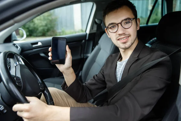 Young man is sitting at his car while looking at the camera. His left hand is on the steering wheel. He is holding the phone in his right hand. — Stock Photo, Image