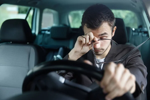 Il giovane assonnato si strofina gli occhi con la mano destra. La sua mano sinistra è sul volante. Lui è seduto alla sua macchina. Concetto sicurezza stradale . — Foto Stock