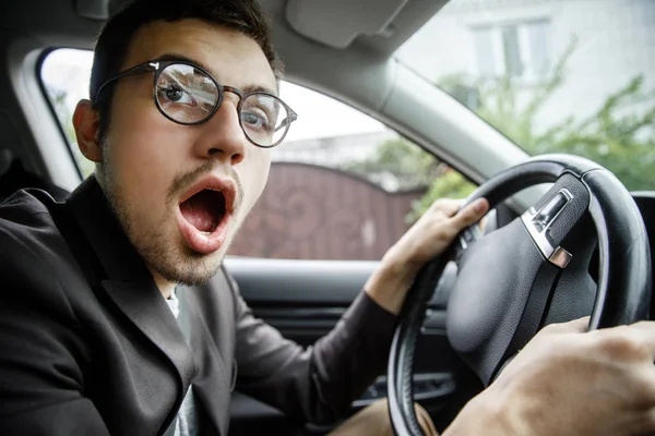 Giovane ragazzo sbadiglia mentre guarda la fotocamera. Lui è seduto alla sua macchina. Ha le mani sul volante. . — Foto Stock