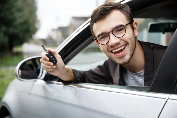 Jovem satisfeito está espreitando da janela do carro enquanto olha para a câmera. Ele está segurando as chaves na mão direita. Conceito vencedor da loteria . — Fotografia de Stock