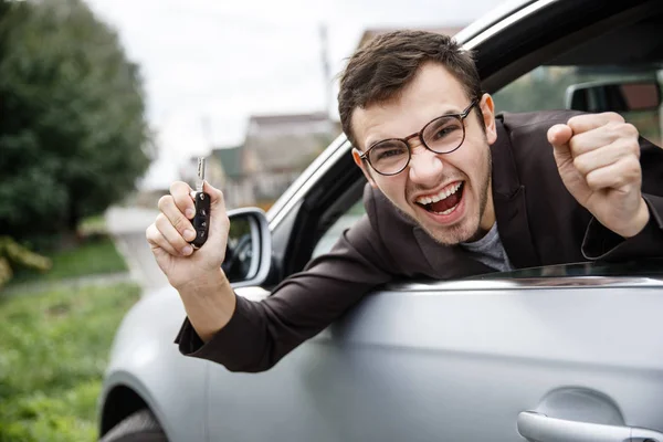 Zachwycony młody człowiek jest peeking z okna samochodu patrząc na kamery. W prawej ręce trzyma klucze. Jego usta są szeroko otwarte. Koncepcja zwycięzca loterii. — Zdjęcie stockowe
