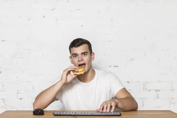 A young guy eats at work on the computer. He's sitting at a table eating a hamburger and typing on his keyboard. — Stock Photo, Image