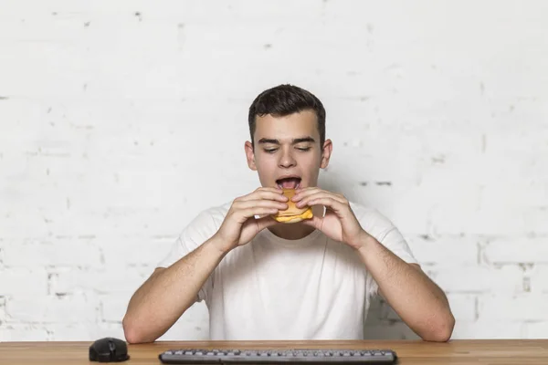 A young man eats at the workplace. He eats a hamburger. On the table there is a keyboard and mouse. — Stock Photo, Image