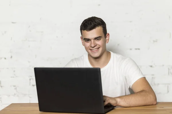 Bir masada oturan ve bir dizüstü bilgisayar üzerinde çalışan genç bir adam. Gülümsüyor.