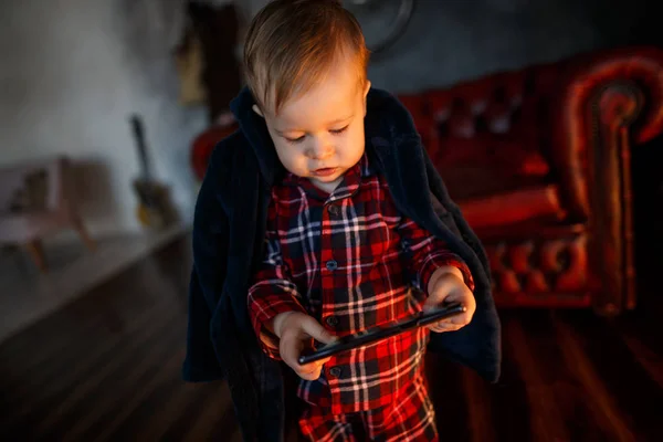 Small baby is standing and holding tablet in hands. He is looking at it. Child is concentrated. He is in room near red sofa.