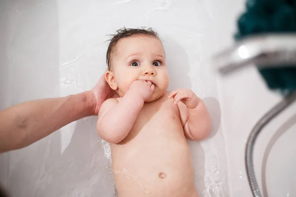 Nice and beautiful baby is lying in bath and looking up. She is naked. Baby is holding her small fist in mouth. Baby's head is holded by adult's hand. Isolated on white background. — Stock Photo, Image