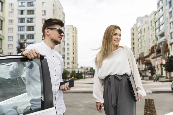 Mooi paar staan samen buiten op straat. Guy is leunend op auto's deur. Hij draagt een bril. Meisje kijkt naar dezelfde richting. Ze houdt van computer in handen. — Stockfoto