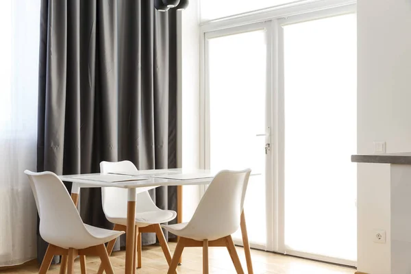 Neben dem großen weißen Fenster stehen ein weißer Holztisch und drei Plastikstühle. schwarzer Vorhang auf der Rückseite. — Stockfoto