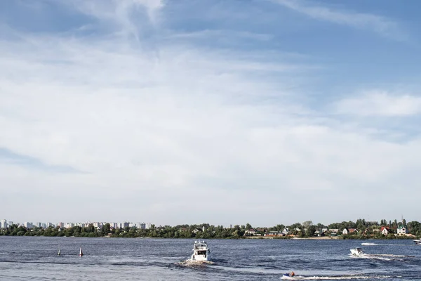 Big river with some boats and a big pleasure boat sailing in it. Big modern city far at the horizon line.