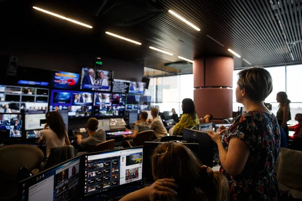 Çalışan bir sürü kişi ile geniş yayın merkezi. Arka planda monitörlerle kaplı bir duvar.