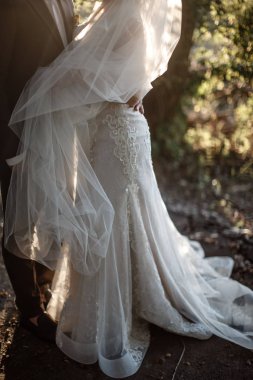 Bir damat ve gelin ormanda sarılıyorlar. Gelin beyaz bir gelinlik giyiyor ve bir damat siyah bir takım elbise giyiyor.