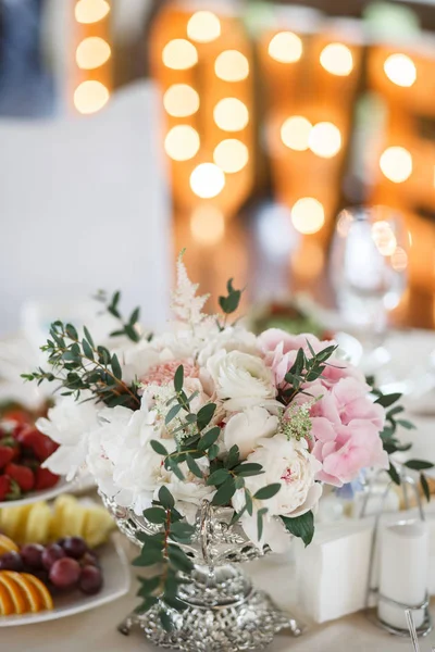 A flower bouquet in a silver pot is standig on a table alongside with some plates with food. Blurred decorations at the background.