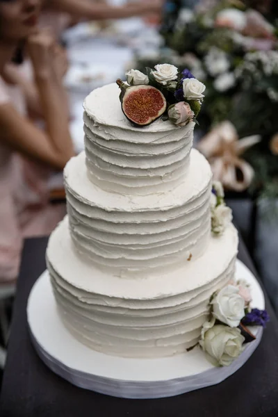 A big wedding cake, decorated with the flowers and a fruit. Table with a guests sitting behind it is at the background.