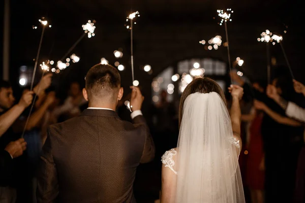 A bride and a groom are holding a burning sparklers up in the air. A lot of guests are holding their sparklers at the background. — Stock Photo, Image