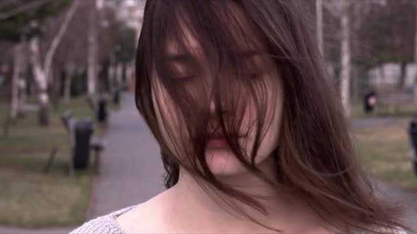 En vacker flicka står i parken, vinden är waing hennes hår. Hon tittar på kameran, sedan blundar och blickar ner. — Stockvideo