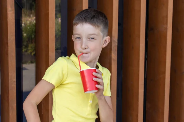 一个穿着黄衬衫的男孩站在木墙边 拿着一只红色的一次性纸杯 用吸管喝水 — 图库照片