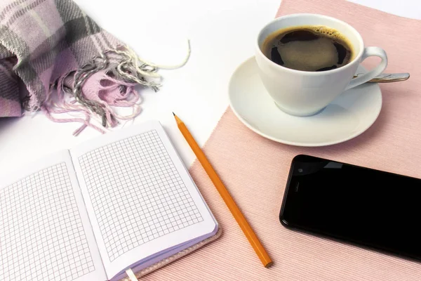 White ceramic cup of coffee, notebook, pencil and smartphone on white table, covered with pink cloth and woolen scarf.