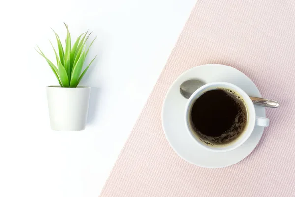 White ceramic cup of coffee and green fake plant in white pot on white table, covered with pink cloth.