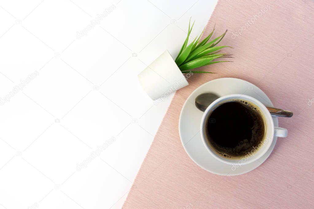 White ceramic cup of coffee and green fake plant in white pot on white table, covered with pink cloth with copy space.
