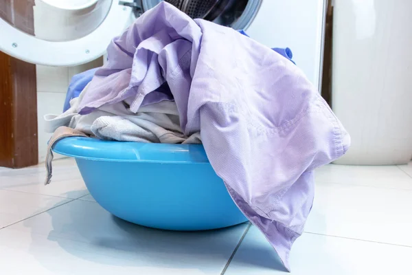 A basin full of dirty laundry on the bathroom floor, blurred washing machine on background.