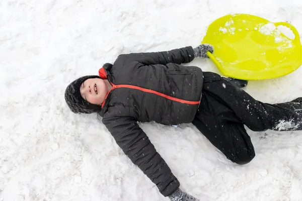 一个7岁的男孩躺在雪上 手里拿着绿色的塑料雪橇 冬季活动 娱乐和儿童娱乐的概念 — 图库照片