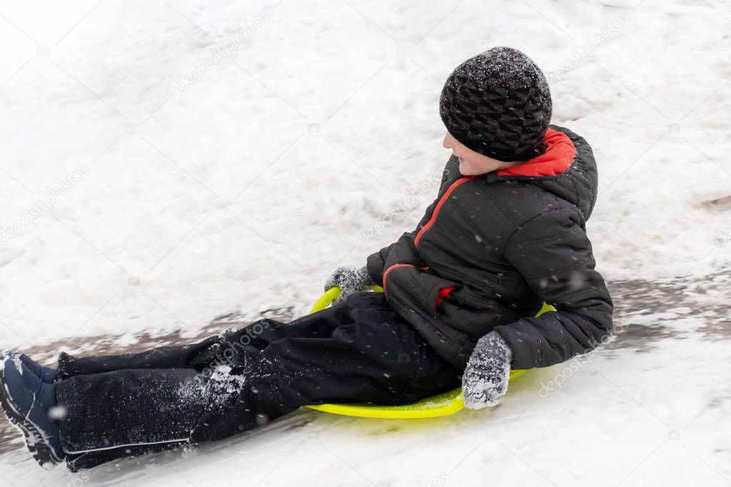 A boy of seven years old rides the slide , down the hill on green ice sled. Concept of winter activities, recreation and children's entertainment.