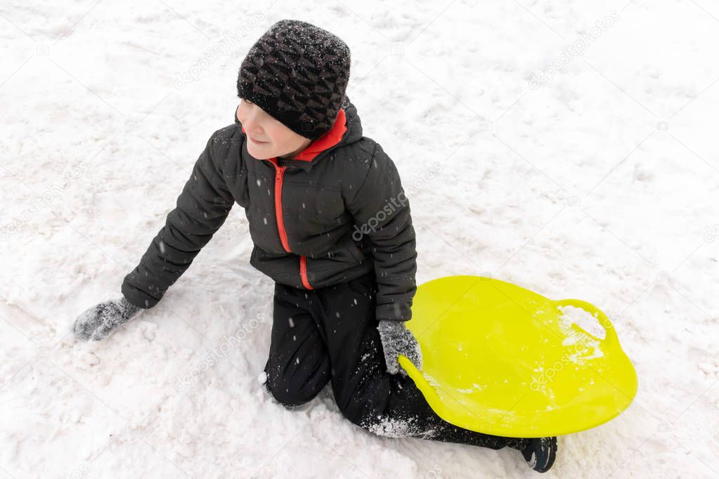 A boy of seven years old lying on the snow and holding a green plastic sled in his hand. Concept of winter activities, recreation and children's entertainment.
