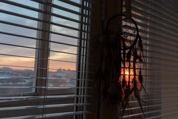 Handmade dreamcatcher hanging by the window with blinds in sunset twilight. Black silhouette of traditional magic amulet for dream protection and bad spirits.